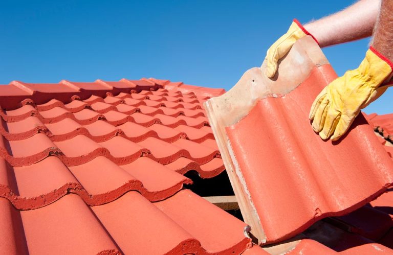 A gloved worker placing a red terracotta roof tile onto a tiled roof under a clear blue sky.